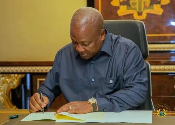 President John Dramani Mahama is pictured at a desk in an official setting, focused on signing a document. The Coat of Arms of Ghana is visible in the background on a wooden panel, signifying a formal state action at Jubilee House.
