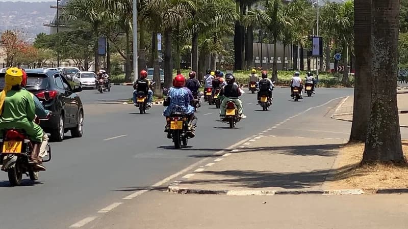 A photograph showing a fleet of commercial motorcyclists, commonly known as motos, navigating an orderly urban road lined with palm trees. Each rider and passenger is visible wearing a helmet, demonstrating the disciplined and regulated nature of Rwanda's motorcycle transport system.