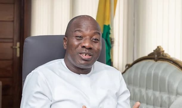 A medium shot of Ghanaian politician Mahama Ayariga speaking in an indoor office setting. He is wearing a traditional white outfit and is seated in a grey chair, with the Ghana flag and a piece of furniture visible in the background.
