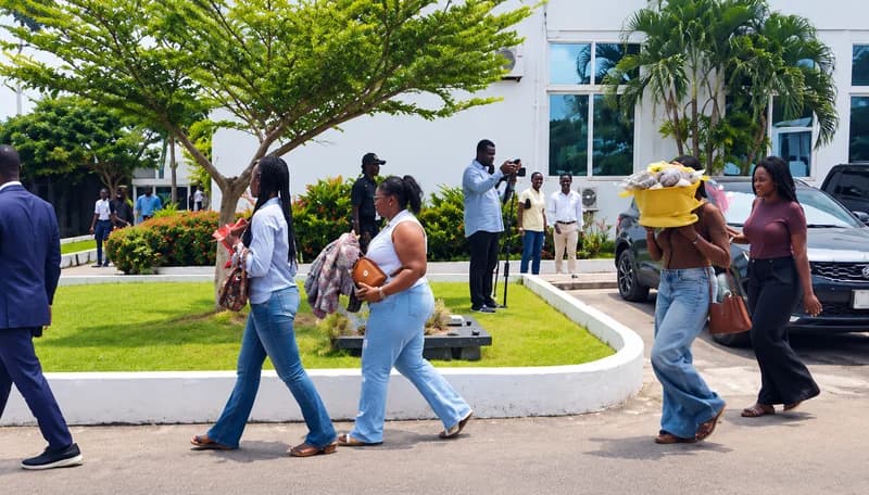 Several women are seen walking through a paved courtyard outside a white building, likely after being apprehended. One woman is carrying a large yellow money bouquet on her head, which serves as evidence in the case involving the illegal use of Ghanaian cedi notes. Observers and a man filming the scene are also visible in the background.