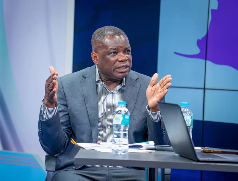 Ghanaian politician Governs Kwame Agbodza gestures emphatically while speaking in a studio setting. He is seated at a desk with a laptop, documents, and water bottles, likely addressing concerns regarding coastal road infrastructure and property demolitions in Osu.