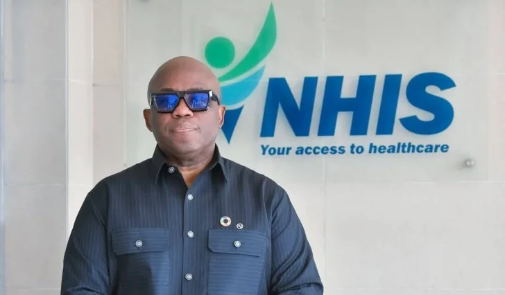 A photograph of Dr. Da-Costa Aboagye, the Chief Executive of the National Health Insurance Authority (NHIA), standing in front of the official NHIS logo. He is wearing a black button-down shirt and blue-tinted sunglasses while positioned against a wall featuring the slogan 'Your access to healthcare'.