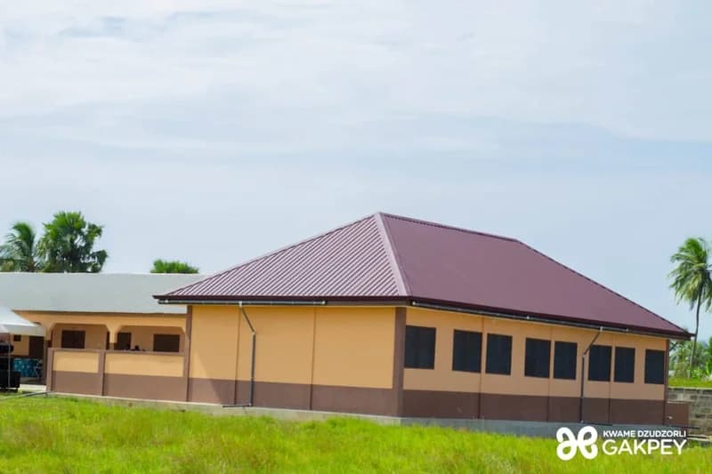 A newly constructed school building, identified as a kindergarten block, featuring a maroon metal roof and beige and brown walls. The structure is situated in an open grassy area with palm trees and a light sky in the background, representing a new educational infrastructure project.