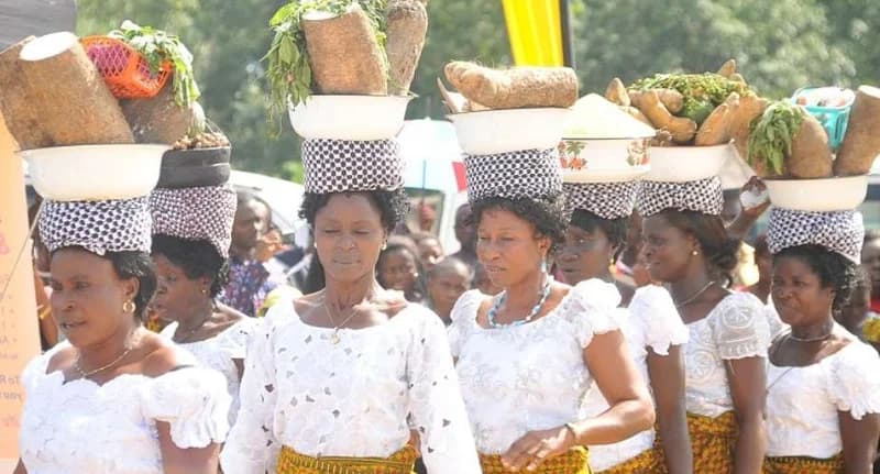 A group of Ghanaian women are seen processing during a traditional festival. They are wearing white lace tops and carry large yams in basins balanced on their heads using checkered headpads. The scene depicts the cultural celebration of a Yam Festival, likely in the Nchiraa area.