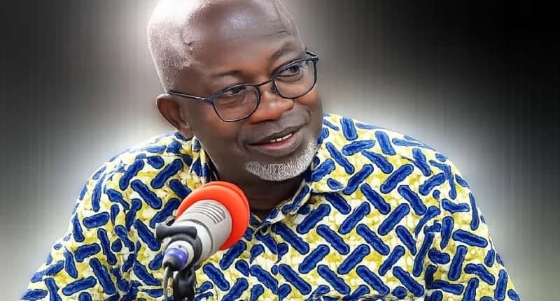 The image features Dr. Richard Amoako Baah, a prominent Ghanaian political scientist, during a media appearance. He is wearing a distinct yellow and blue patterned shirt and speaking into a microphone with a red windscreen. The setting appears to be a studio interview where he is offering political commentary.