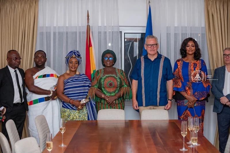 A group of seven individuals, including Ghanaian officials and European representatives, stand together behind a large wooden table in an indoor setting. The scene includes the Ghana national flag and a blue flag, likely the European Union flag, indicating a diplomatic or official event. Several glasses are placed on the table, suggesting a toast or celebratory gathering for the launch of the European Film Festival Ghana 2026.