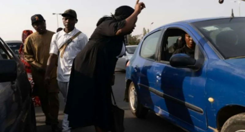 A woman in a black dress gestures emphatically towards a driver in a blue car on a busy road. Several men stand nearby, including one in traditional attire, depicting a scene of potential tension or public frustration often seen during periods of fuel scarcity.