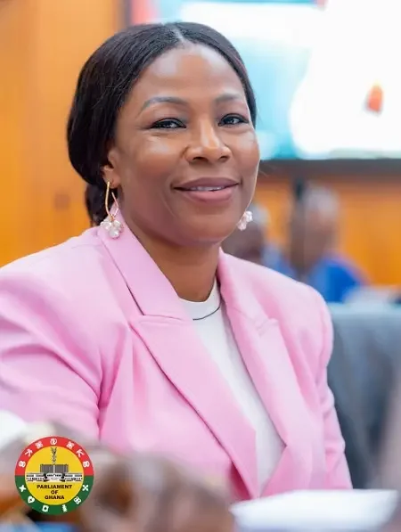 A close-up photograph of a woman, likely a Ghanaian public official or Member of Parliament, wearing a pink blazer and pearl earrings. The official logo of the Parliament of Ghana is visible in the bottom-left corner, and the subject is smiling in an indoor setting with other figures blurred in the background.