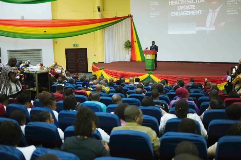 A formal health sector event held at the Ghana College of Physicians and Surgeons, featuring a government official speaking from a podium decorated in Ghana's national colors. A large audience is seated in blue chairs, observing a screen presentation titled 'Health Sector Update Engagement' which highlights progress in Ghana's health systems.
