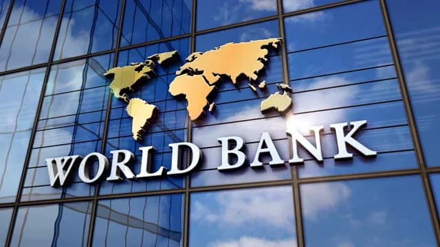 A high-quality digital illustration of the World Bank logo and name prominently displayed on the glass exterior of a modern skyscraper. The 3D silver text and golden world map are set against a background reflecting a clear blue sky with white clouds.