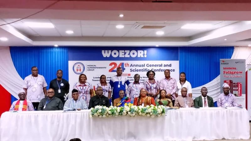 A group of medical professionals and dignitaries pose for a photograph at the 24th Annual General and Scientific Conference of the Medical Superintendents' Group in Ghana. The participants are seated and standing behind a long, flower-decorated head table against a backdrop featuring the conference theme and the Ewe welcome word 'WOEZOR!'.