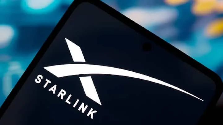 A close-up photograph of a smartphone screen displaying the white Starlink logo and name against a deep black background. The phone is tilted diagonally, and the background consists of a soft-focus bokeh effect with blue and teal highlights.