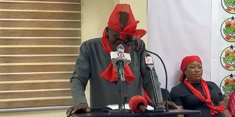 A man wearing a red headpiece and scarf speaks at a podium during a press conference or protest. A woman in similar red attire is seated behind him, suggesting a collective show of mourning or grievance common in Ghanaian public demonstrations.
