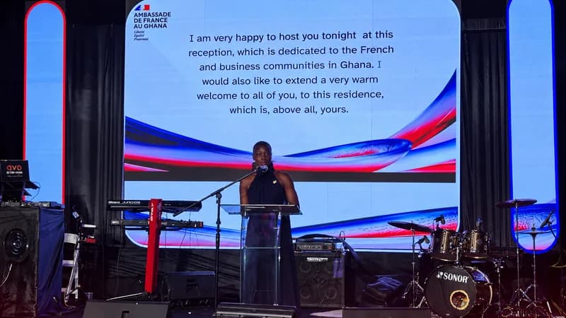 A woman stands at a transparent podium delivering a speech during a formal diplomatic event. Behind her, a large screen displays the French Embassy logo and a welcoming message addressed to the French and business communities in Ghana. The setting includes musical equipment like a drum kit and keyboard, suggesting a reception or gala atmosphere.