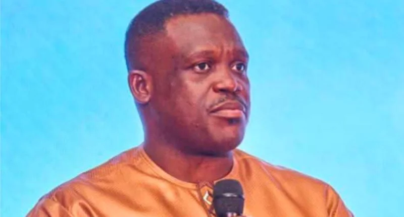 A close-up photograph of Samuel Nartey George, a prominent Ghanaian politician, speaking into a microphone. He is wearing a copper-toned traditional tunic against a vibrant blue studio background, appearing focused and serious during an interview or address.