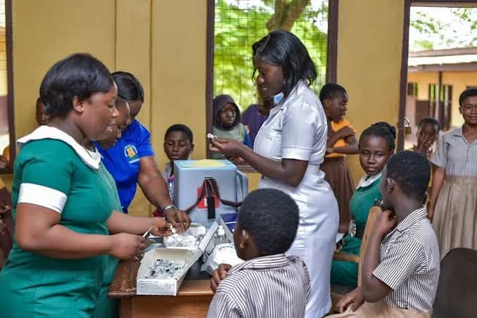 A team of healthcare workers, including nurses in green and white uniforms, are seen preparing medical supplies and vaccine vials at a desk. They are surrounded by schoolgirls in uniforms, suggesting a vaccination exercise taking place within a Ghanaian school setting.
