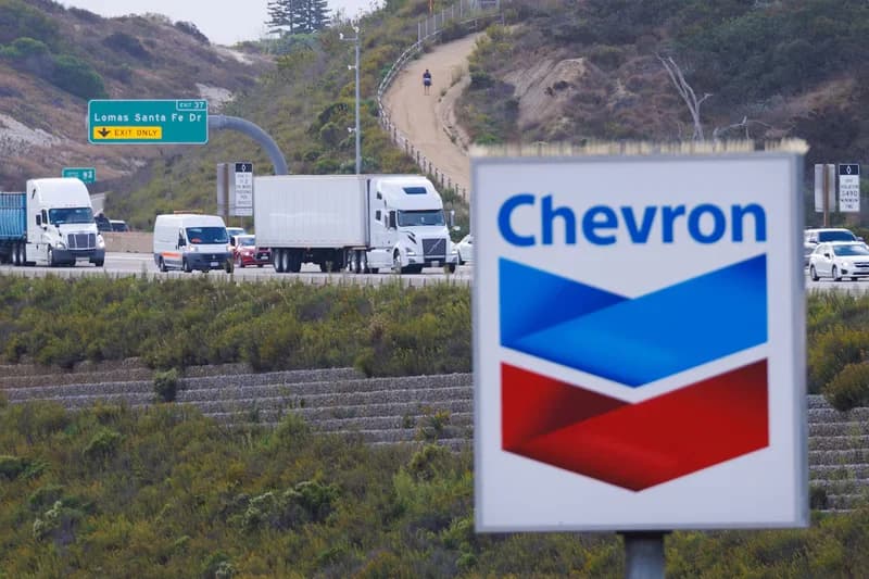 A prominent Chevron corporate logo sign is positioned in the foreground of a highway scene. Several large white semi-trucks and a van are visible on the road behind it, with a green exit sign for Lomas Santa Fe Drive and a coastal hill in the background.