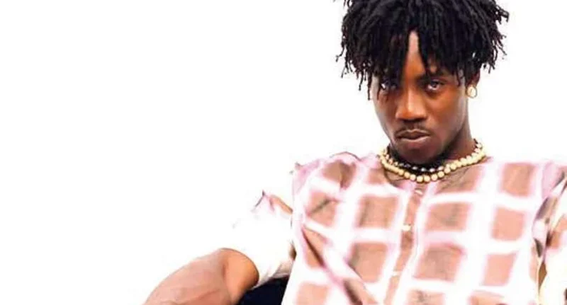 A portrait of Ghanaian artist AratheJay featuring him in a checkered patterned shirt and a double-strand beaded necklace. He is looking directly into the camera against a plain white background, likely as a promotional image for his upcoming concert.