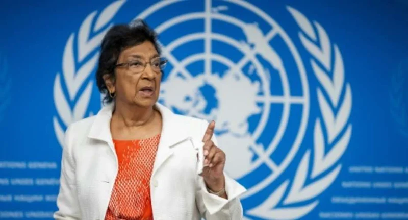 Navi Pillay, the South African jurist and human rights activist, is seen speaking at a podium with her finger raised. She is wearing a white blazer over an orange patterned shirt, standing against a blue backdrop featuring the United Nations logo.