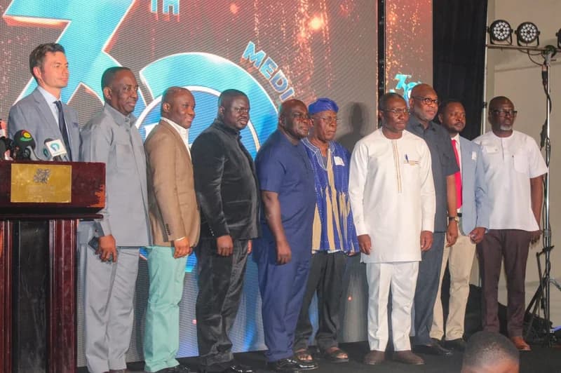 A group of prominent men, including media practitioners and officials, stand together on a stage at a formal event in Ghana. Behind them is a large digital backdrop displaying a "30th" anniversary logo with the word "MEDIA" partially visible. To the left, a podium with multiple microphones suggests a series of speeches or a press conference is taking place.