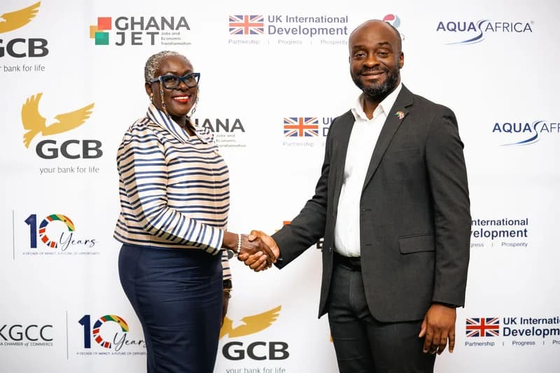 A man and a woman share a professional handshake in front of a media backdrop featuring various organizational logos. Both are dressed in formal business attire and are smiling toward the camera during what appears to be a ceremonial or corporate event.