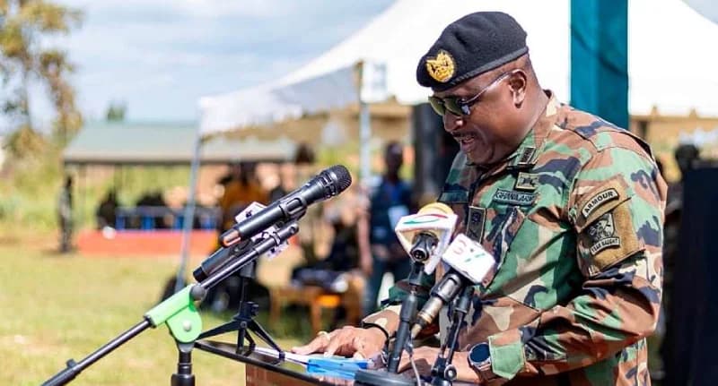 A high-ranking Ghana Army officer in camouflage uniform and a black beret is seen speaking at a podium equipped with several microphones. He is wearing sunglasses and looking down at his speech notes during an outdoor military event, with white tents and other personnel visible in the background.