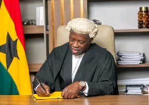 The image depicts Emmanuel Armah-Kofi Buah dressed in traditional legal attire, including a black gown and a white barrister's wig. He is seated at a wooden desk, focused on signing a document with a yellow cover, while the Ghana national flag is visible in the background. This photograph captures the moment following his formal enrollment as a lawyer in Ghana.