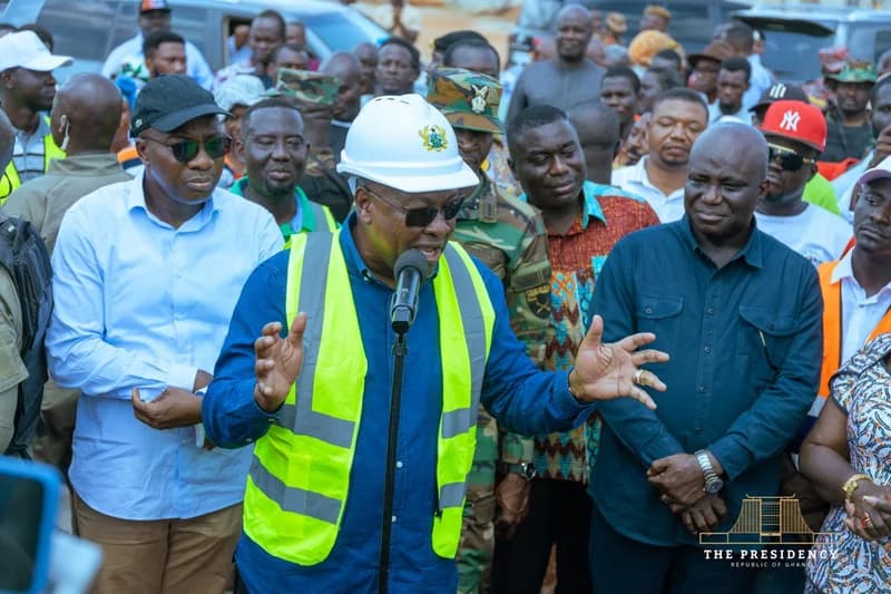 President John Dramani Mahama wears a white hard hat and a high-visibility safety vest while speaking at a project site inspection. He is surrounded by a diverse group of people, including security personnel in camouflage and various officials, as part of his regional project tour.