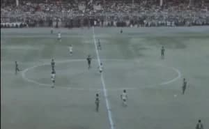 A wide-angle, low-resolution archival photograph showing a football match in progress at a crowded stadium. Players are distributed across the pitch around the center circle, with a dense crowd of spectators visible in the background stands.
