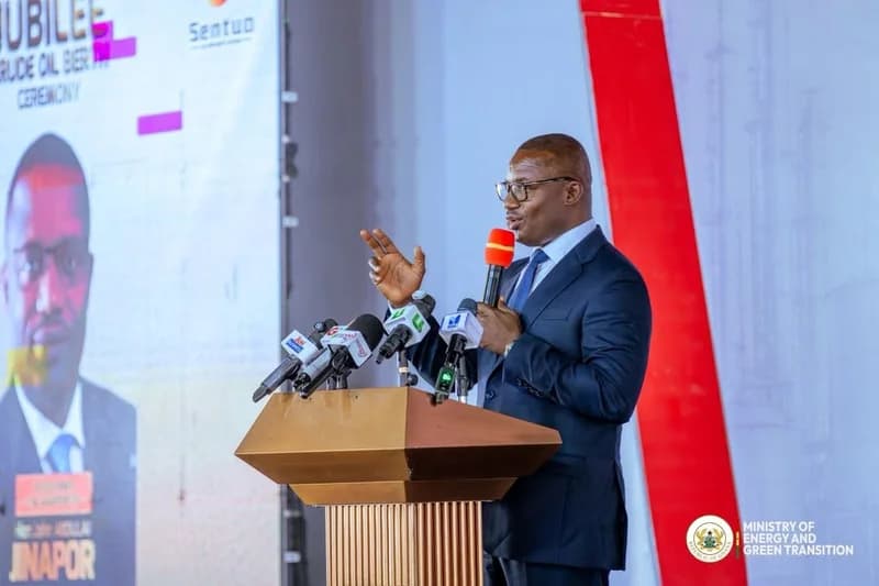 Hon. John Jinapor, Ghana's Minister of Energy and Green Transition, is pictured speaking at a podium during an official event. He is gesturing with one hand while holding a microphone, surrounded by several media microphones, with a background banner detailing a Jubilee Crude Oil Berth ceremony.