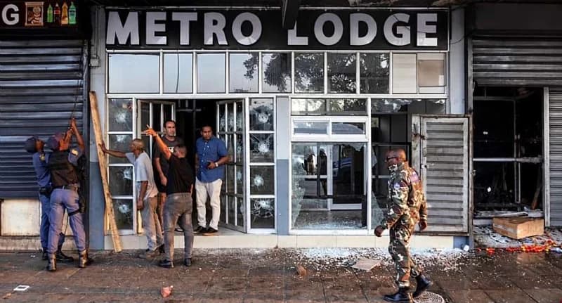 The image shows the vandalized storefront of 'Metro Lodge' with several shattered glass windows and visible projectile holes. South African police officers and several men are seen inspecting the damage outside the building where glass shards litter the pavement.
