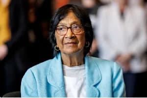 A professional portrait of Navi Pillay, the South African jurist and former UN Human Rights chief. She is seen wearing a light blue blazer, glasses, and gold hoop earrings, looking calmly off-camera with other figures blurred in the background.