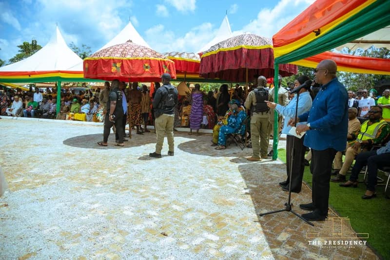 President John Mahama is captured speaking at a microphone during an outdoor commissioning ceremony in Ghana. The event features traditional ceremonial umbrellas, large white marquees decorated with national colors, and a gathering of officials, security personnel, and citizens.