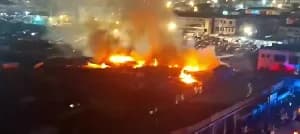 A large fire consumes several market stalls at night, sending bright orange flames and thick smoke into the air. In the background, a crowd of onlookers observes the blaze under the glow of streetlights in an urban setting.