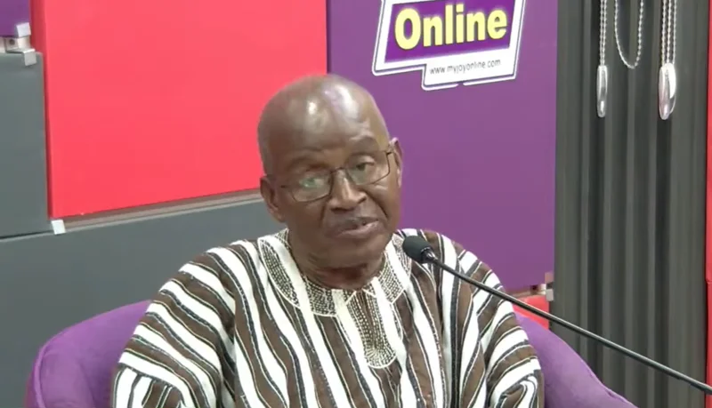 Retired judge Justice Francis Korbieh is captured in a studio setting, wearing a traditional striped Ghanaian smock (fugu) and glasses while speaking into a microphone. The background features red and purple panels with a Joy Online logo, suggesting a media interview or presentation.