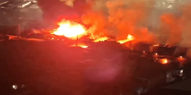 A large, intense fire engulfs market structures at night, with bright orange flames and thick smoke rising into the air. The scene depicts a major fire outbreak, likely the one reported at the Kumasi Central Market.