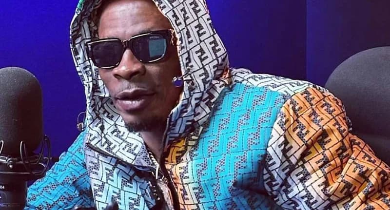 A photograph of Ghanaian dancehall musician Shatta Wale in a studio setting. He is wearing a vibrant, multi-colored patterned hooded jacket and dark sunglasses, seated in front of a professional microphone.