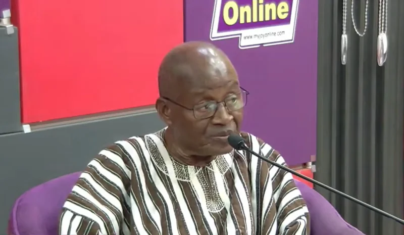 Justice Francis Korbieh is captured during a media interview, wearing a traditional Ghanaian smock and speaking into a microphone. The background features a purple and red studio setting with a prominent Joy Online logo visible behind him.