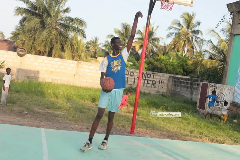 Ephraim Saawa, a 7-foot tall Ghanaian, stands on an outdoor basketball court holding a basketball and reaching toward the hoop. The photograph captures him in his sporting environment at Ngleshie Amanfro, with palm trees and a wall featuring the words 'DO NOT LITTER' in the background.