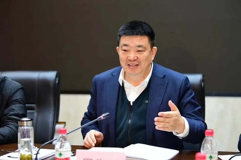 A photograph of former Wuhan mayor Zhou Xianwang speaking at a meeting. He is dressed in a dark blue blazer and a white shirt, gesturing with his hand while seated at a table equipped with a microphone and water bottles.