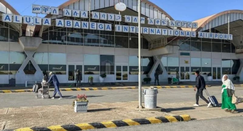The image shows the exterior of the Alula Aba Nega International Airport terminal in Mekelle, Ethiopia. Travelers with luggage and carts are visible in the foreground under a clear blue sky, with the airport's name displayed in large English and Amharic letters.