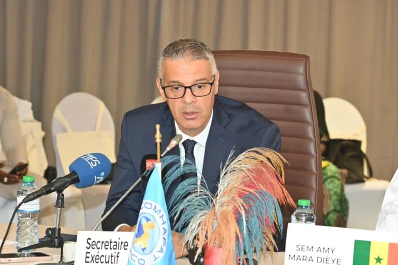 A man in a dark suit and glasses sits at a conference table in Senegal. He is positioned behind microphones and a nameplate reading 'Secretaire Exécutif', with a COMHAFAT flag and a Senegal flag visible on the table, indicating a formal international meeting.