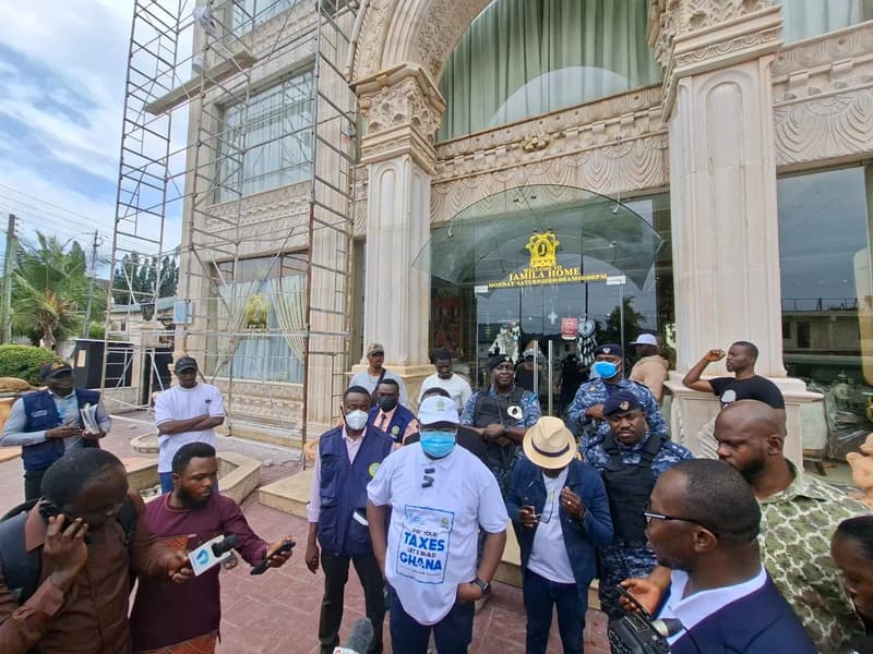 Officials from the Ghana Revenue Authority (GRA) accompanied by security personnel are seen during an enforcement exercise at the Jamila Home premises. A central official wears a white t-shirt with the slogan 'Pay Your Taxes, Let's Build Ghana', while media personnel record the event in front of the building which is partially covered in scaffolding.