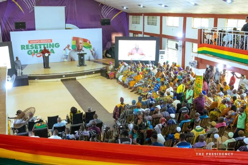 President John Mahama addresses a large audience during the 'Resetting Ghana Tour' in the Ahafo Region. The indoor event features a stage with a prominent banner of Mahama and the Ghanaian flag, with many attendees dressed in traditional Kente and smocks.