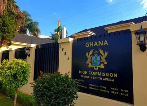 The image displays the entrance and signage of the Ghana High Commission in Pretoria, South Africa. A prominent black sign features the Ghanaian coat of arms along with office hours and location details, situated next to a cream-colored wall and gated perimeter.