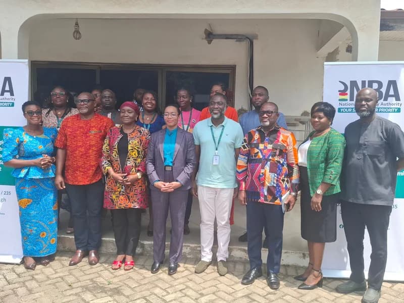 A group of officials and representatives from the National Biosafety Authority (NBA) pose for a group photograph in front of a building in Ghana. The individuals are dressed in a mix of formal Western suits and colorful traditional African prints, flanked by two branded roll-up banners.