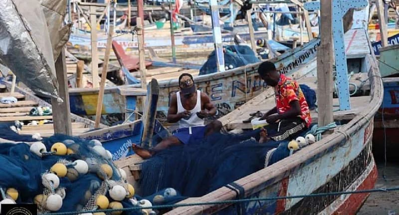 Two fishermen are seen sitting inside a traditional wooden canoe, meticulously working on large blue fishing nets equipped with yellow and white floats. The background shows a dense collection of similar artisanal fishing boats docked at a harbor, representing a typical Ghanaian fishing community.
