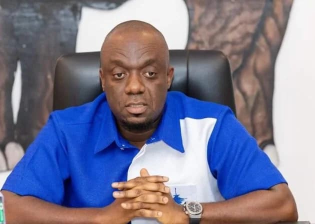 A medium-shot photograph of Justin Kodua Frimpong, the General Secretary of the New Patriotic Party (NPP), seated at a desk with his hands clasped. He is wearing a blue and white shirt reflecting party colors against a background that includes a portion of the NPP elephant logo.