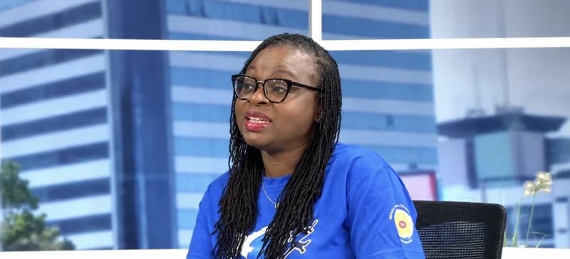A woman with long black braids and glasses is speaking during a studio interview. She is wearing a blue t-shirt with a white graphic and is positioned against a background featuring a blurred urban cityscape.