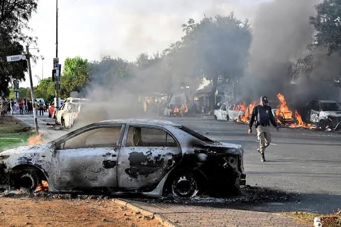 A street scene showing several cars set on fire and emitting thick black smoke during a period of civil unrest. A uniformed security officer walks past a charred vehicle in the foreground while a crowd of people gathers in the background. The image captures the aftermath of violent protests, likely related to the anti-migrant incidents described in South Africa.